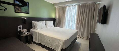 A hotel room with a bed, a desk, a chair, a TV, and a window with curtains.