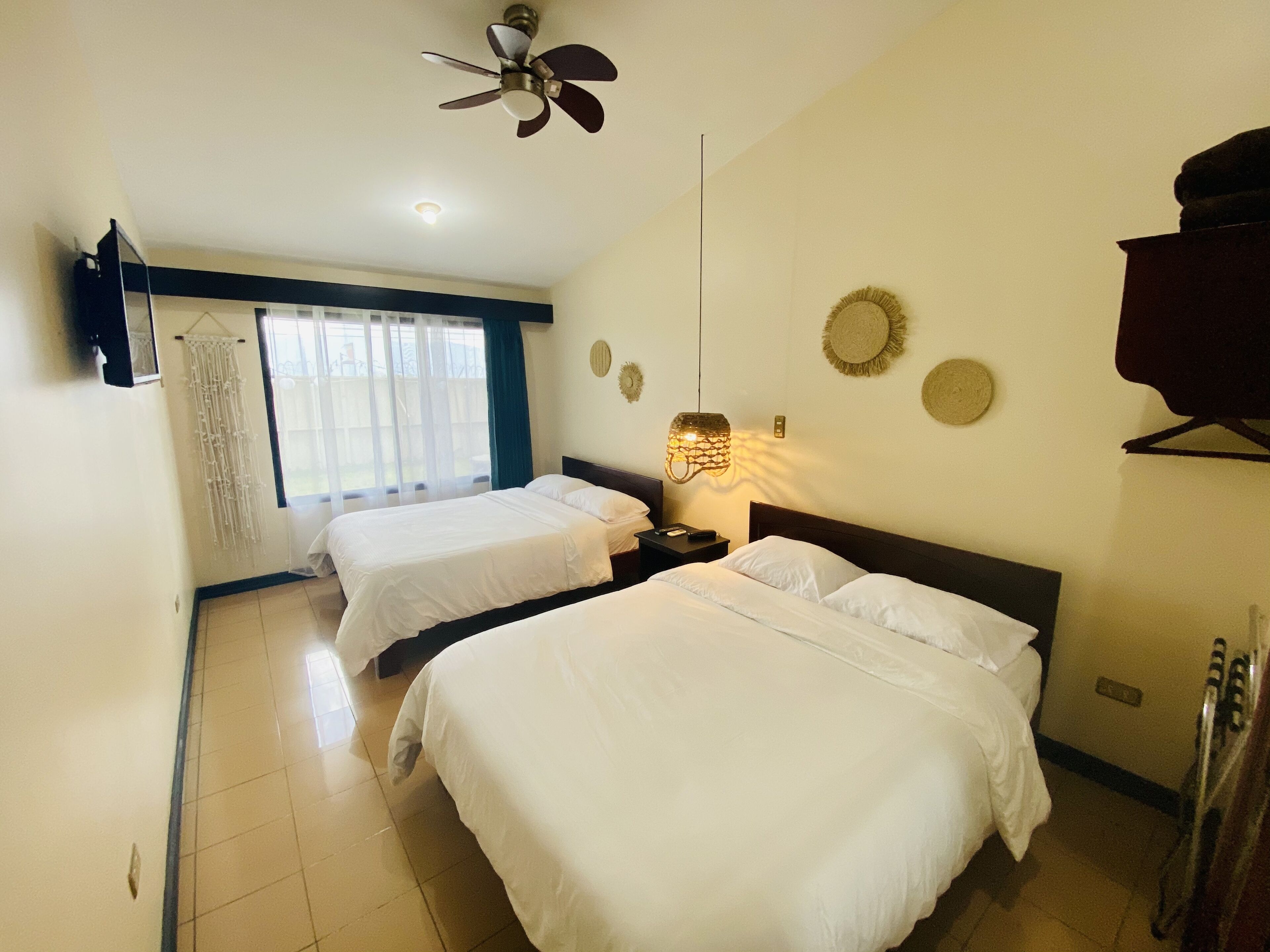 A hotel room with two beds, a ceiling fan, a hanging light fixture, and a view of the outdoors through a window.