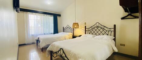 A room with two single beds, a ceiling fan, a bedside lamp, and a window with sheer curtains.
