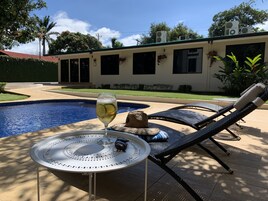 Un área junto a la piscina con una tumbona, una mesa con una bebida en un vaso, y una casa al fondo.