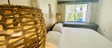 Un dormitorio con cama, una silla de mimbre, un tapiz de macramé y una ventana con cortinas.