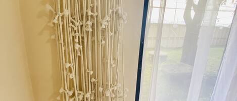 Une décoration murale en macramé dans une pièce donnant sur un jardin, visible à travers des rideaux légers.