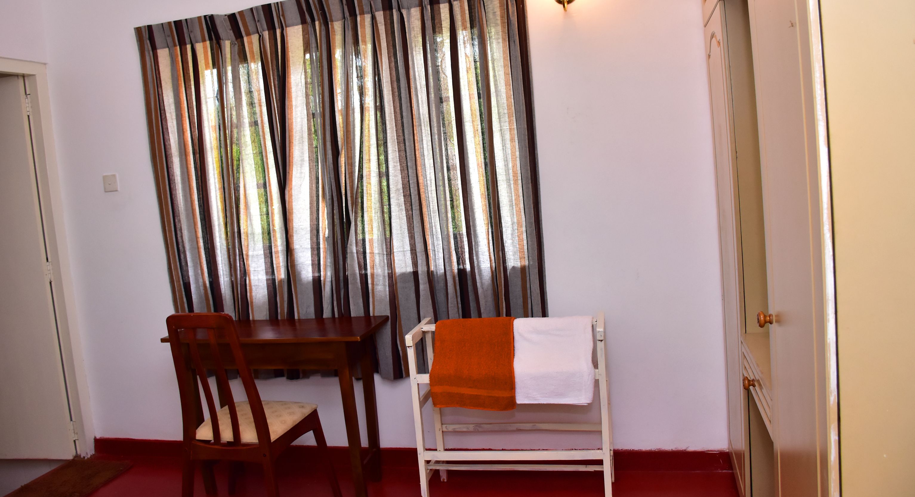 Una habitación con una mesa y silla de madera, un banco blanco con una toalla naranja, y una ventana con cortinas a rayas.
