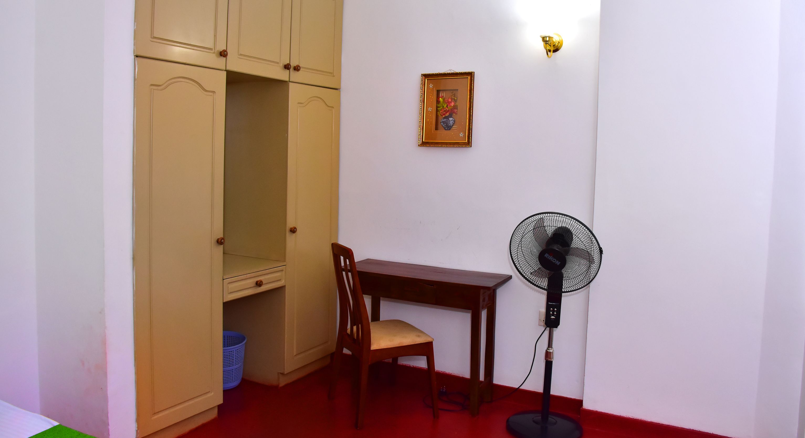 Una habitació amb un armari de fusta, un escriptori, una cadira i un ventilador de peu.