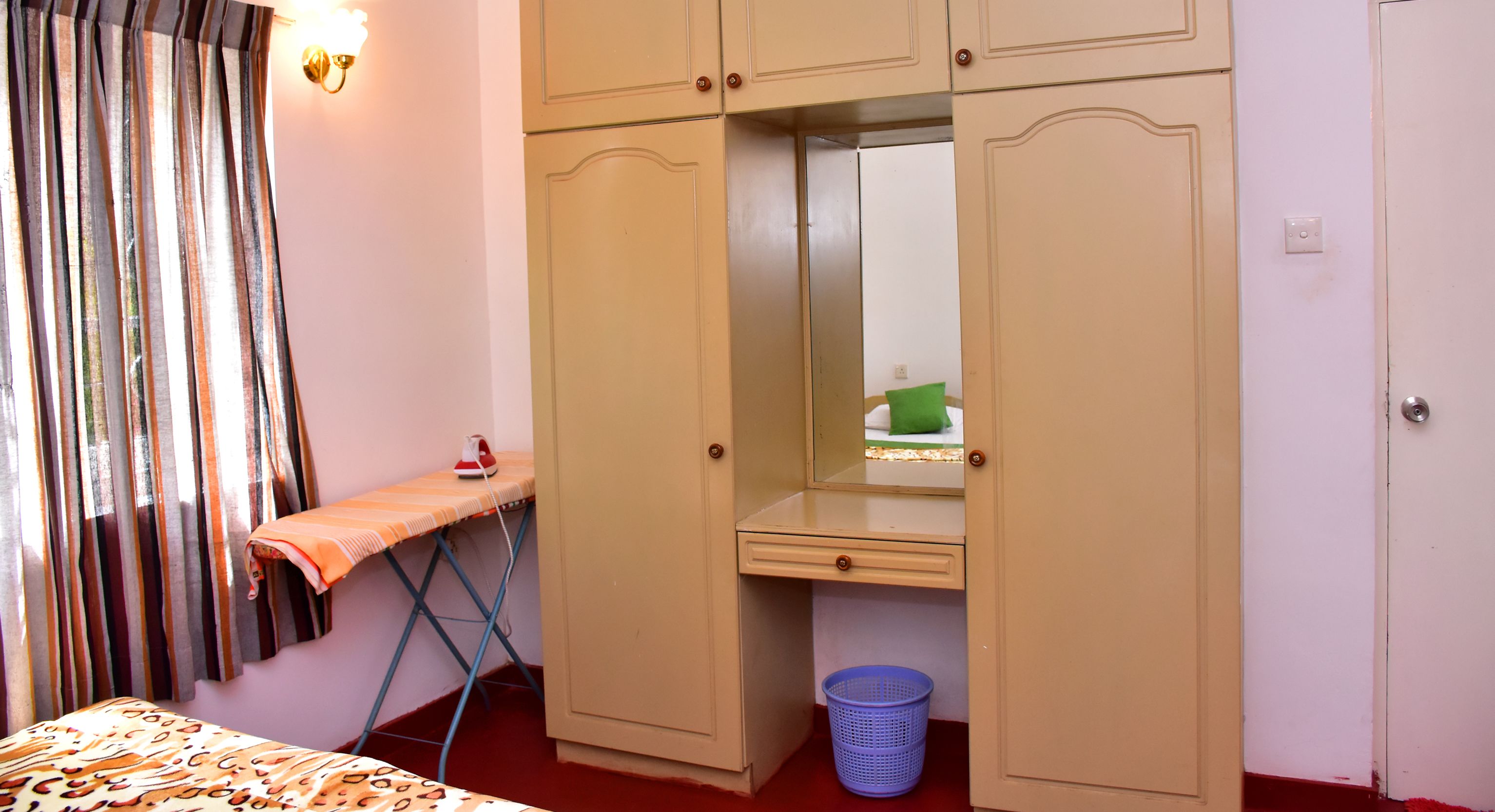 A bedroom with a bed, an ironing board, a wardrobe, a mirror, and a laundry basket.