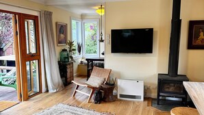 A cozy living room with a fireplace, a television, and a large window with a view of greenery. - Arrowtown Gem - Pet Friendly Arrowtown Cottage. (Arrowtown)