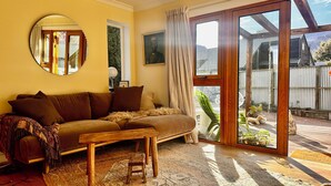 A cozy living room with a sofa, a small wooden table, and a large sliding glass door leading to a garden. - Arrowtown Gem - Pet Friendly Arrowtown Cottage. (Arrowtown)