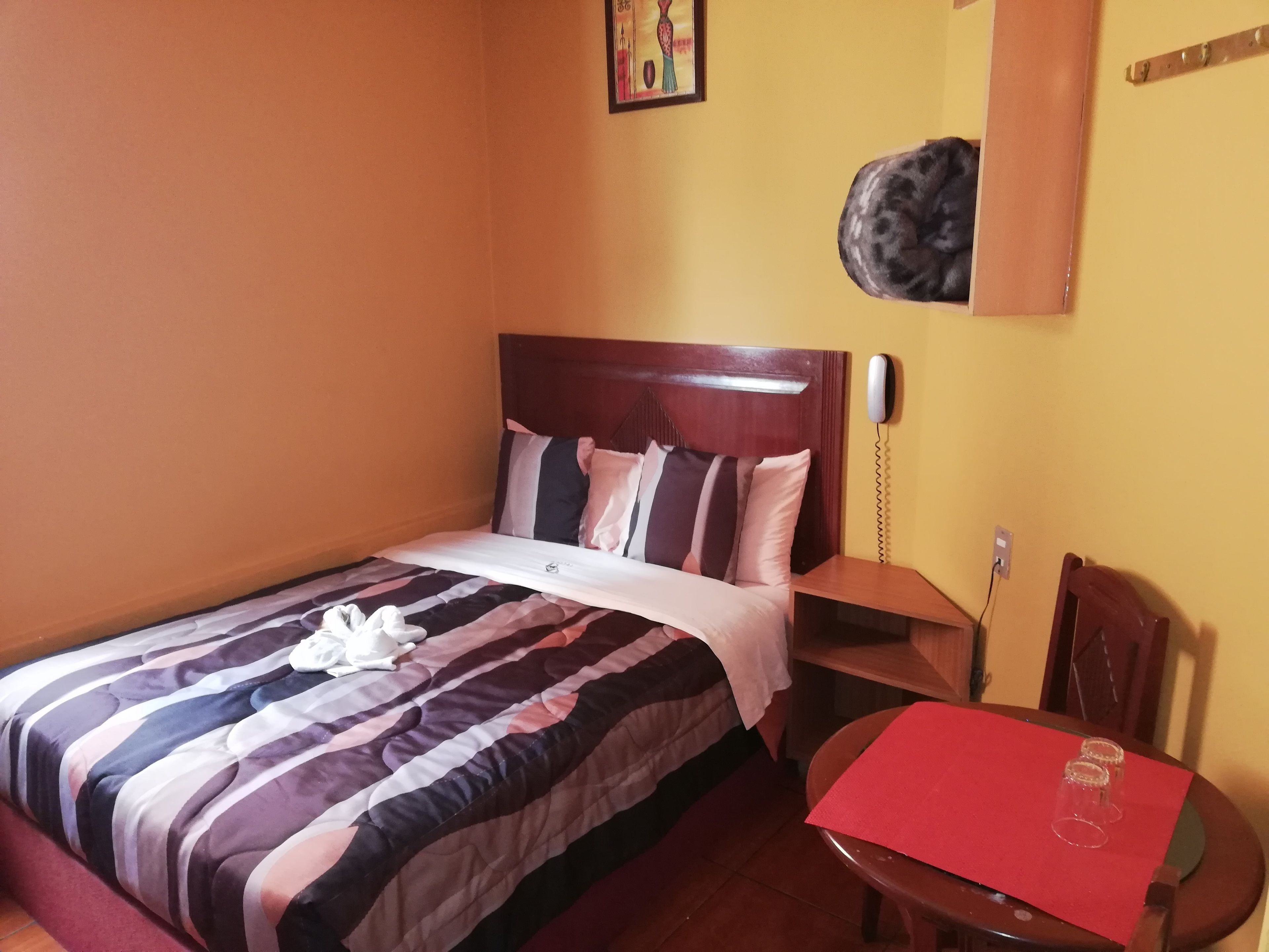 Cama bem arrumada com roupa de cama listrada e um travesseiro, mesa de cabeceira de madeira com um telefone, uma pequena mesa com um jogo americano vermelho e copos, e uma cadeira.