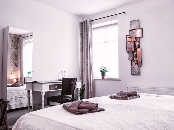 Un dormitorio con cama, escritorio, silla, radiador, ventana con cortinas y una lámpara fijada a la pared.