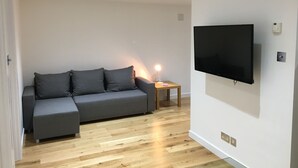 Un salon moderne avec un canapé gris, un téléviseur fixé au mur et une petite table d’appoint en bois sur laquelle se trouve une lampe.