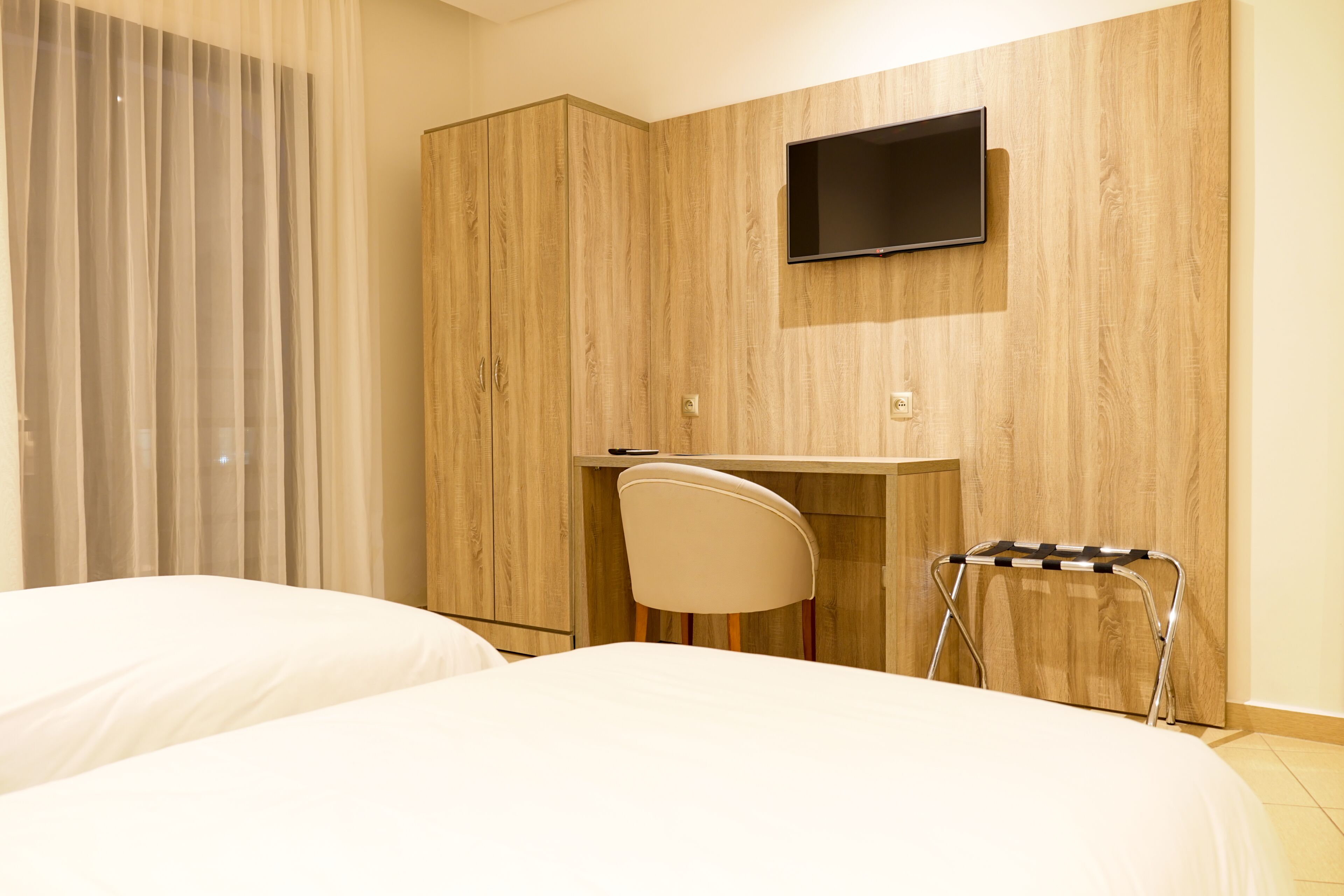 A hotel room with a bed, a desk with a chair, a television mounted on the wall, and a wardrobe.