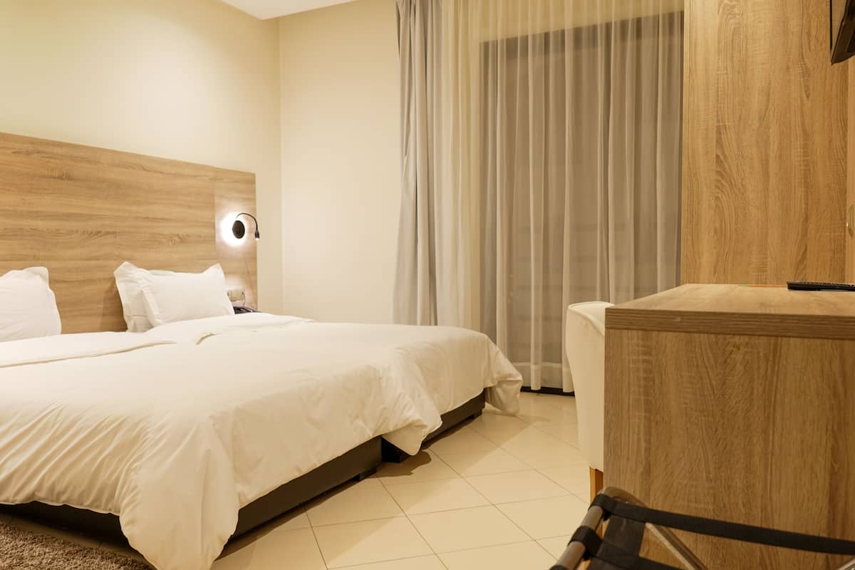 A hotel room with a wooden headboard, a bed with white bedding, a bedside table, and a large window with sheer curtains.