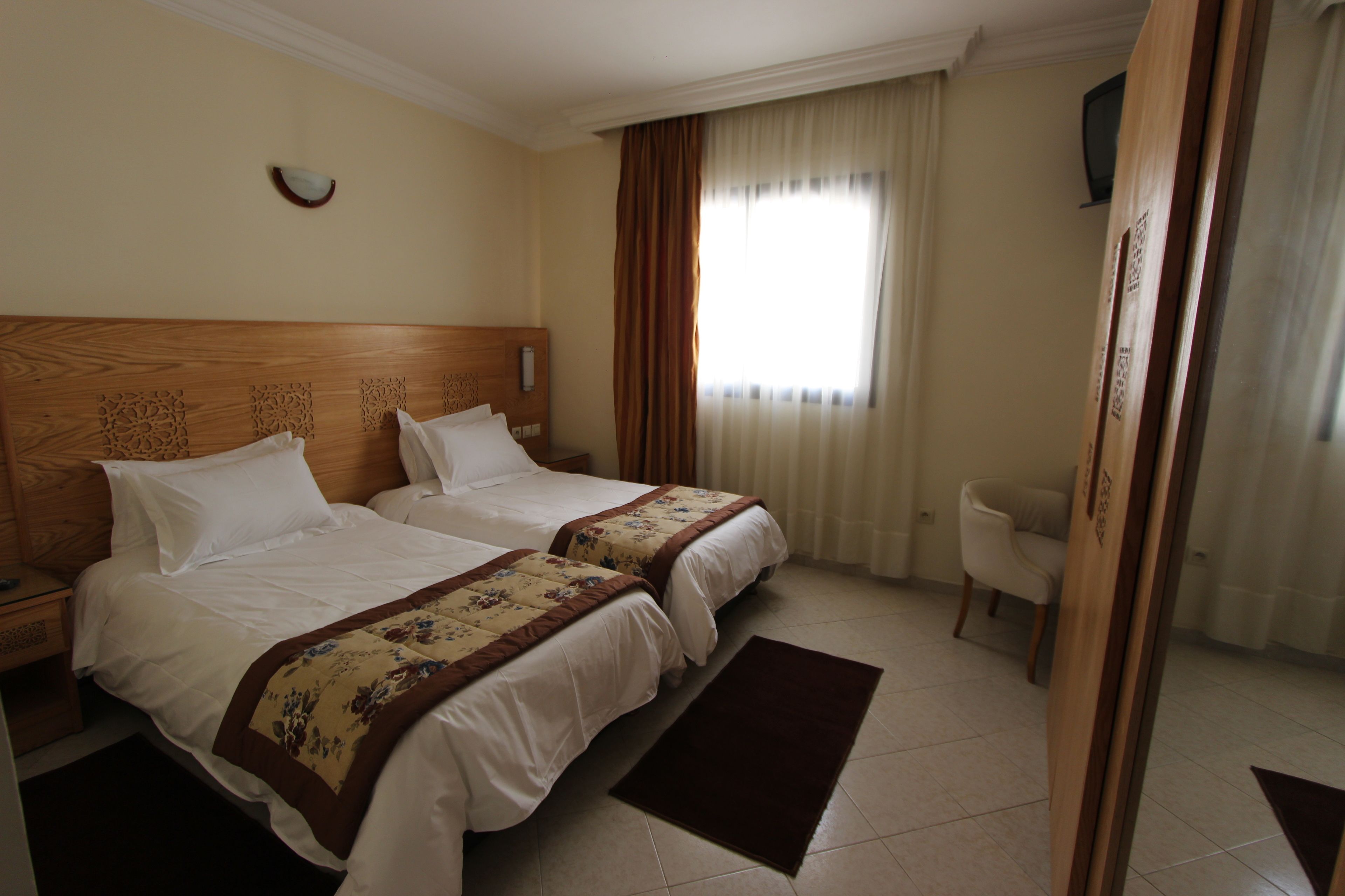 A hotel room with two beds, a wooden headboard, a small desk, a chair, and a television on the wall.