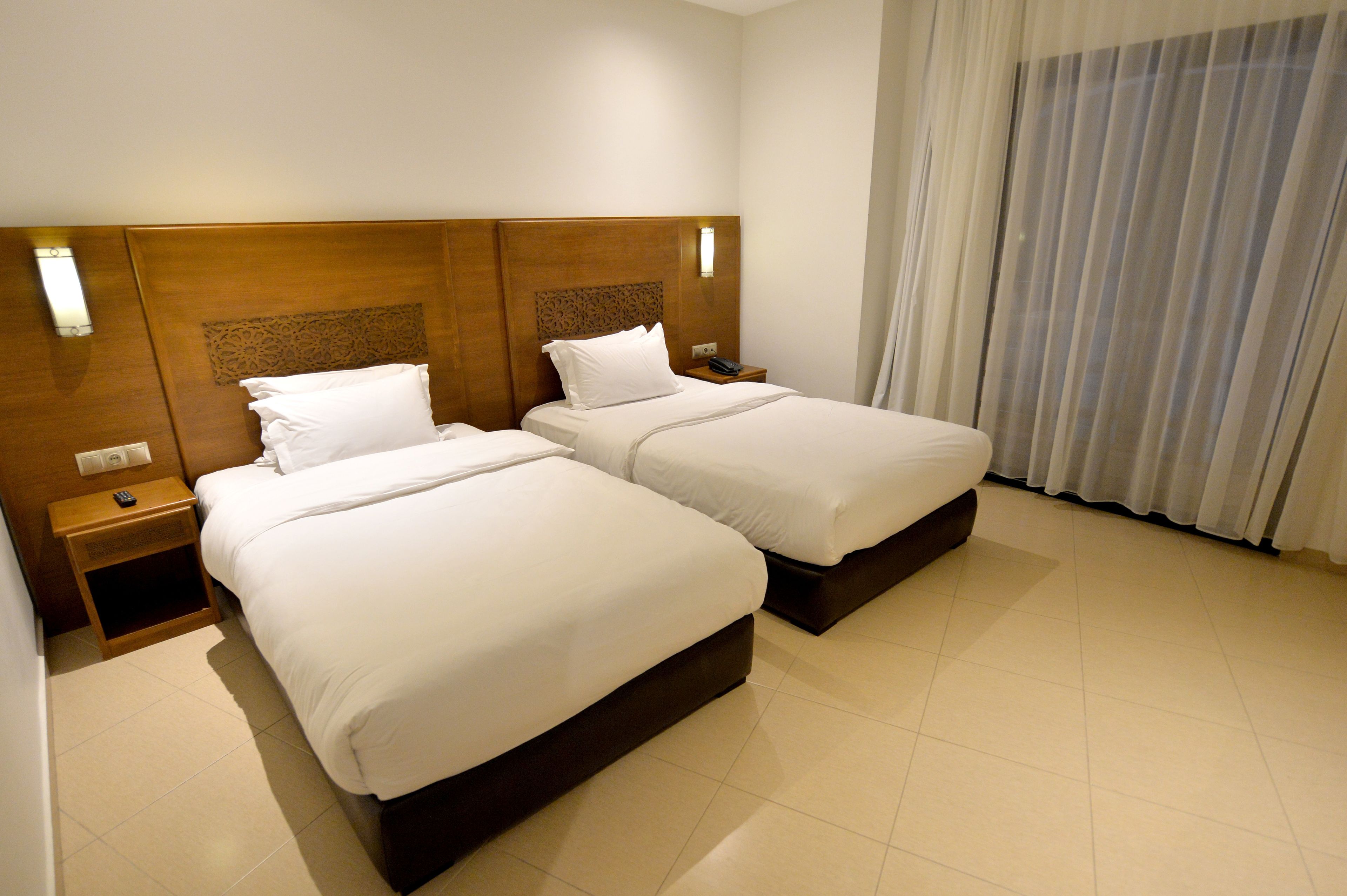 Habitación de hotel con dos camas, cabeceros de madera, mesitas de noche con lámparas y un ventanal con cortinas.