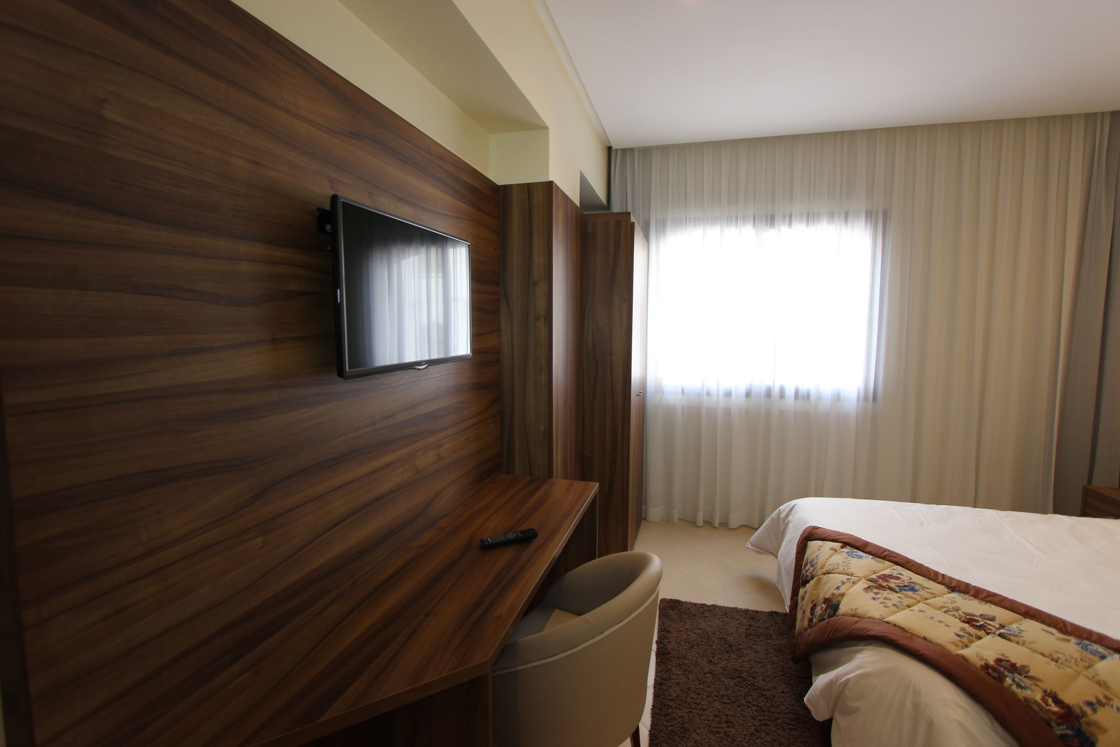 A hotel room with a wooden desk, a flat-screen TV, a bed with a floral bedspread, and a window with curtains.