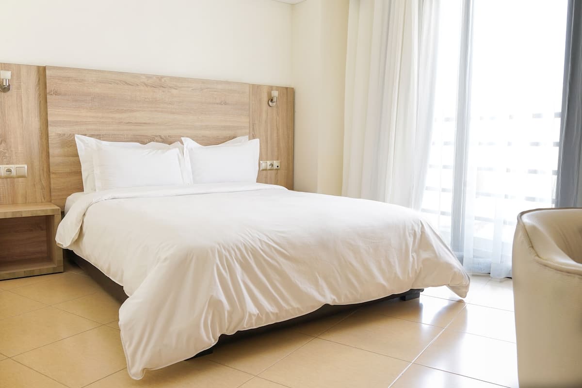 A hotel room with a large bed, a wooden headboard, a nightstand, and a view of the city through the window.