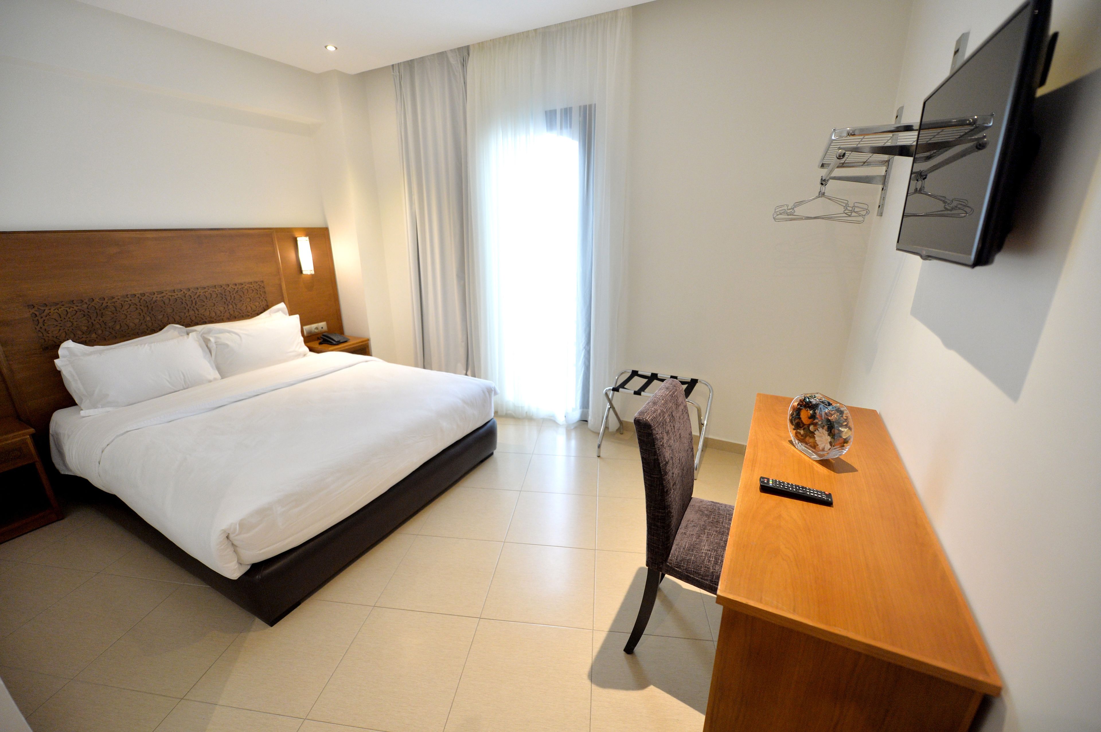 Habitación de hotel con una cama grande, un escritorio con silla, un televisor montado en la pared y una ventana con cortinas.