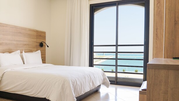 Un dormitorio con una cama grande, cabecera de madera, vista al mar a través de un ventanal amplio y una mesita de noche de madera.