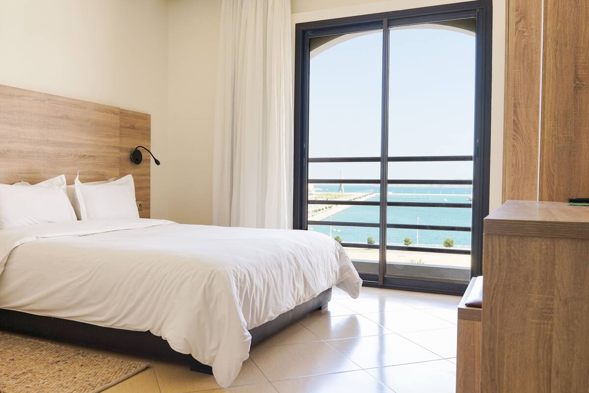 A bedroom with a large bed, a wooden headboard, a view of the sea through a large window, and a wooden nightstand.