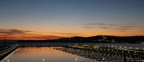 A waterfront marina at dusk with calm water reflecting lights, a pier extending into the water, and a distant hill with lights.