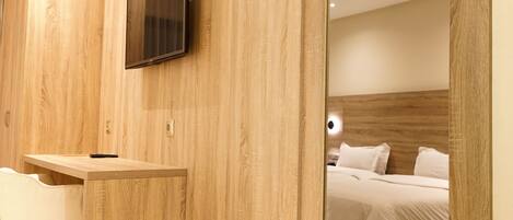 A hotel room with a wooden wall, a mounted TV, a desk with a chair, and a bed with white bedding.
