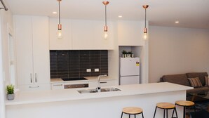 A modern kitchen with white cabinets, a black backsplash, a stainless steel sink, and a built-in refrigerator. There are three bar stools and a small island with stools.
