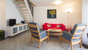 Una sala de estar con un sofá rojo, dos sillones con estampado azul, una mesa de centro, un televisor sobre un mueble bajo y un cuadro de girasoles en la pared.