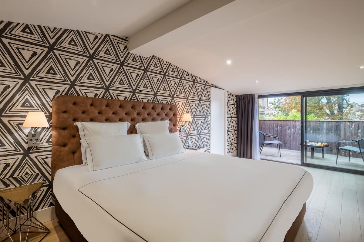 Habitación de hotel moderna con una cama grande, un papel tapiz estampado y un balcón con mobiliario exterior.
