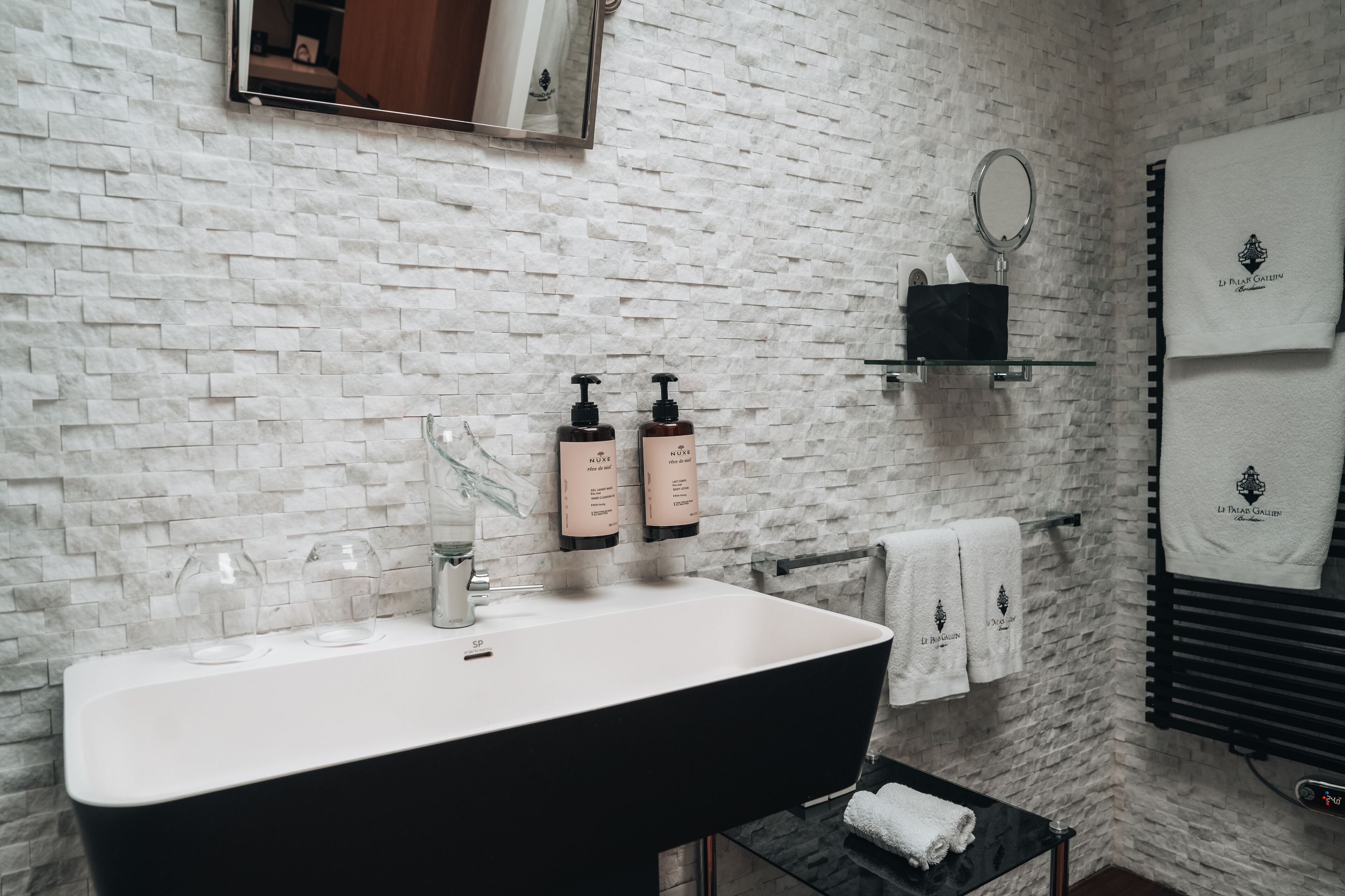 Een badkamer met een witte baksteenmuur, een zwart bad, twee glazen flessen, een spiegel en een handdoekrek met witte handdoeken.