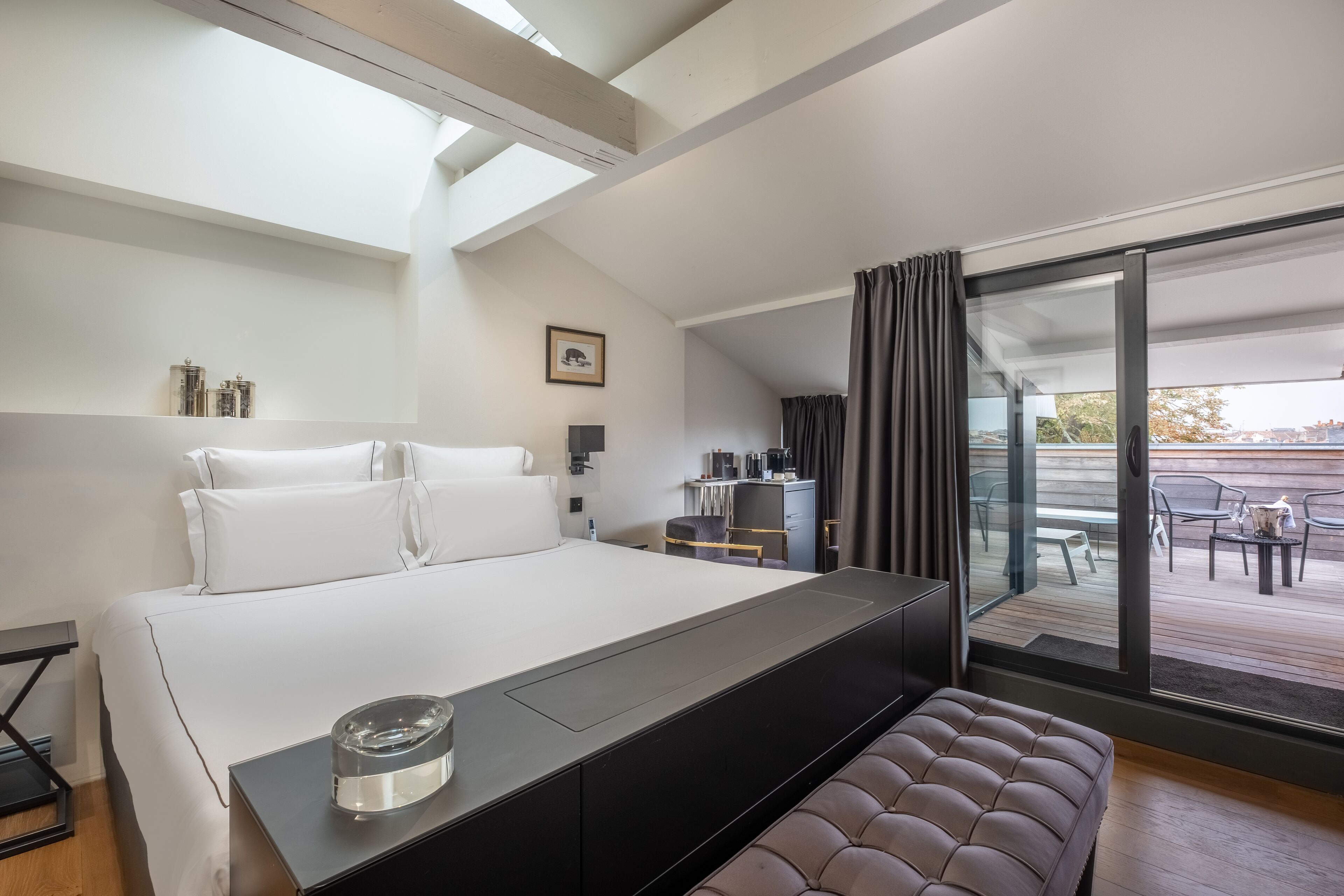 A modern hotel room with a large bed, a bench, a desk with a chair, and a view of the outdoor patio.