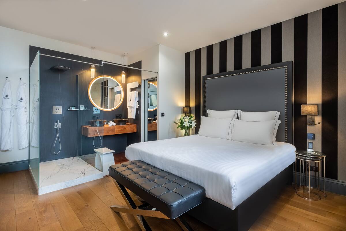 Una habitación de hotel moderna con una cama grande, un baño separado con cabina de ducha de vidrio y una mesita de noche pequeña.