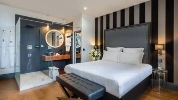 Una habitación de hotel moderna con una cama grande, un baño separado con cabina de ducha de vidrio y una mesita de noche pequeña.