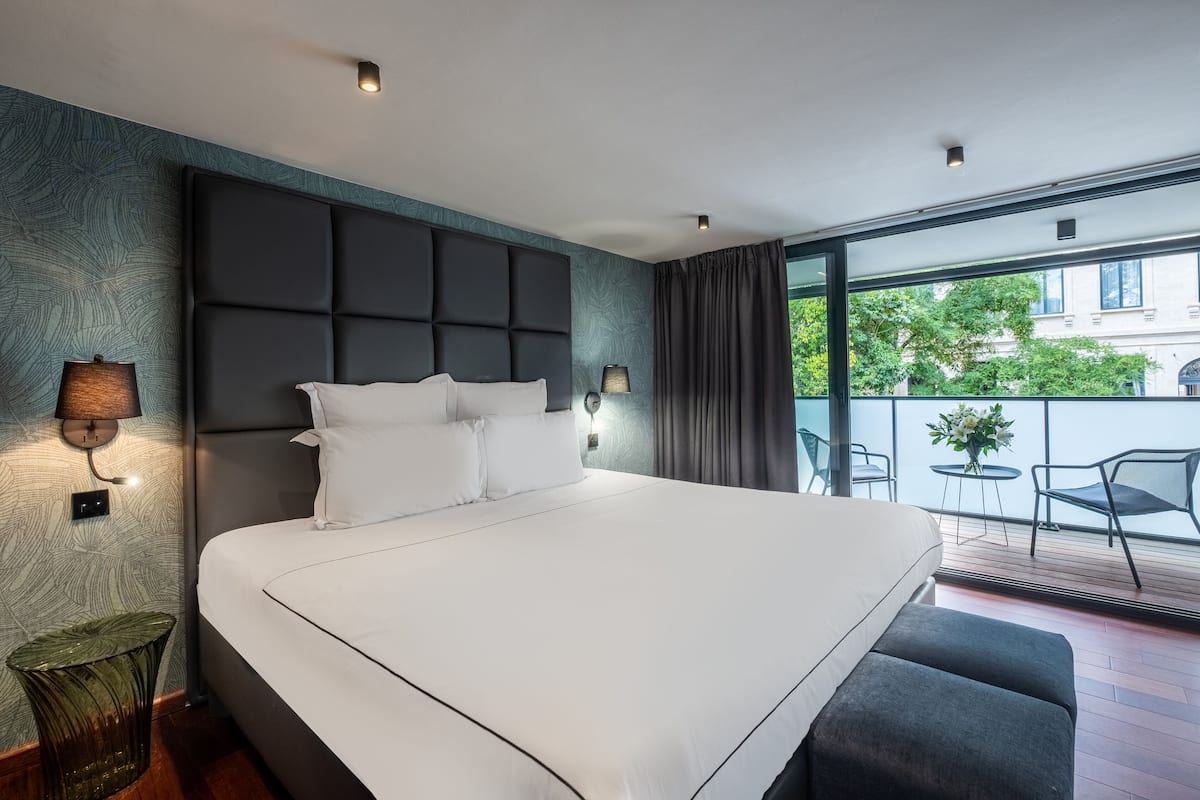 Una habitación de hotel moderna con una cama grande, un balcón con mobiliario exterior y vistas a la ciudad.