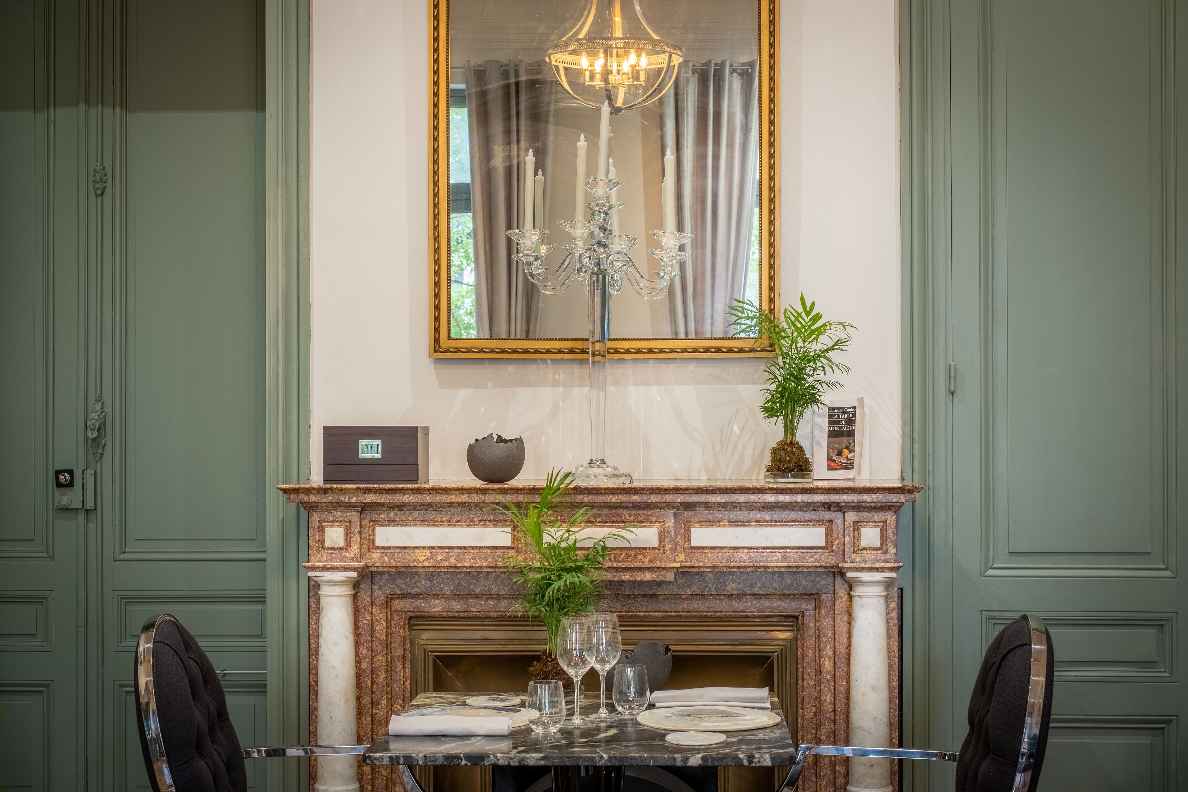 Un comedor con una mesa de mármol, una araña, una chimenea con un espejo encima y puertas verdes.