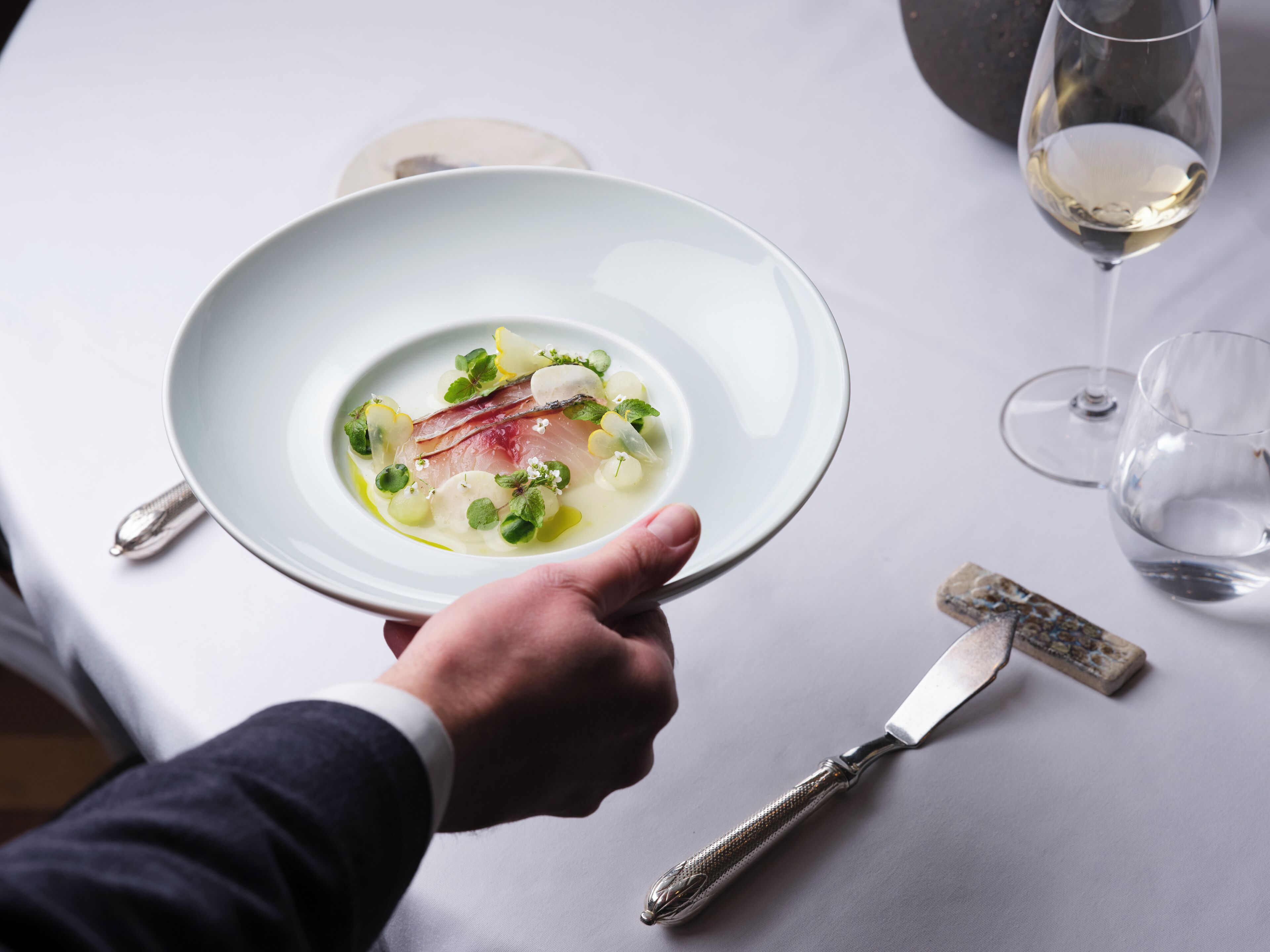 Une personne tenant une assiette blanche sur laquelle se trouve un plat, un couteau et un verre de vin blanc, le tout posé sur une table.