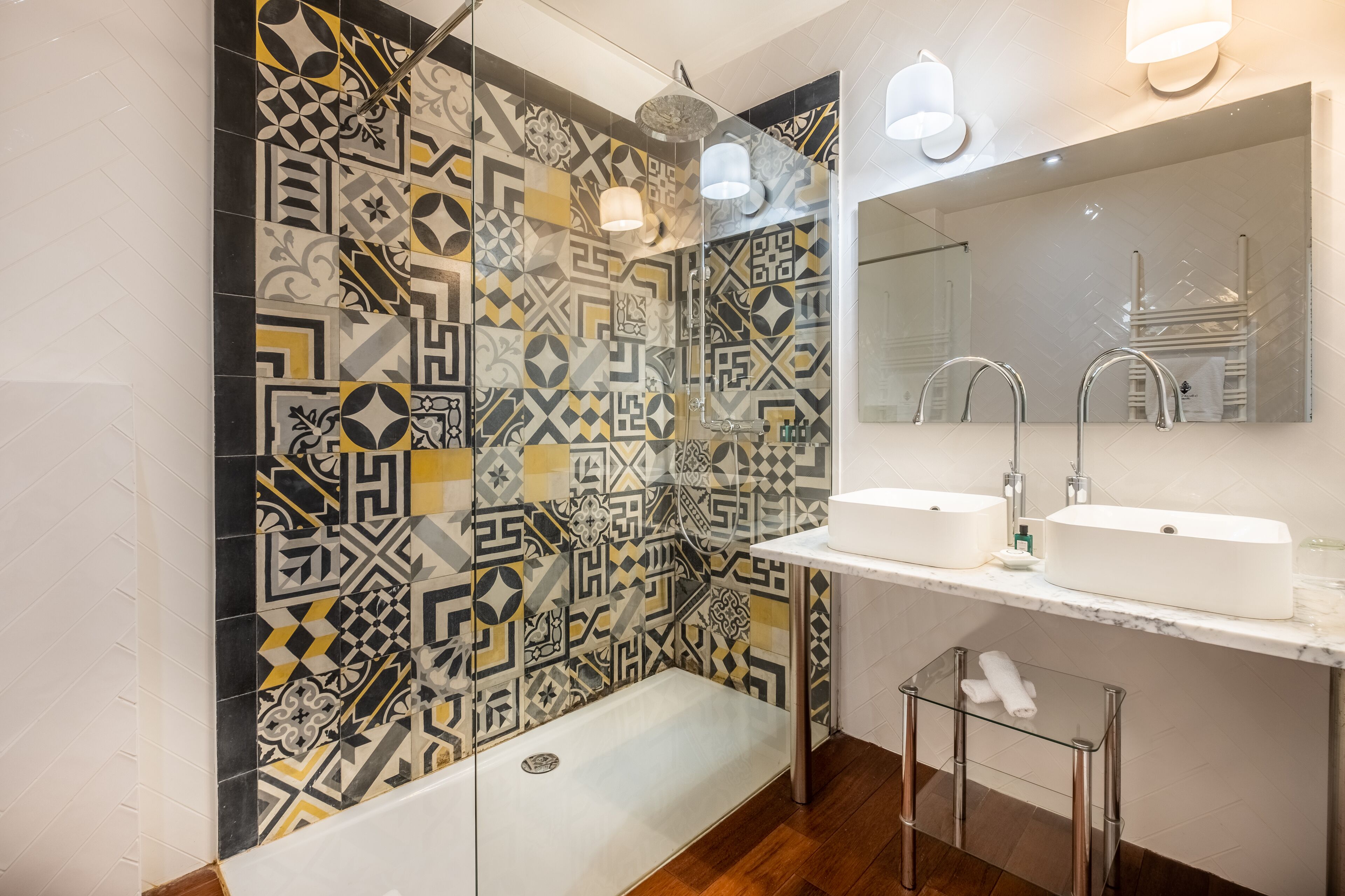 Un baño moderno con baldosas estampadas, cabina de ducha de vidrio, un espejo grande y dos lavamanos.