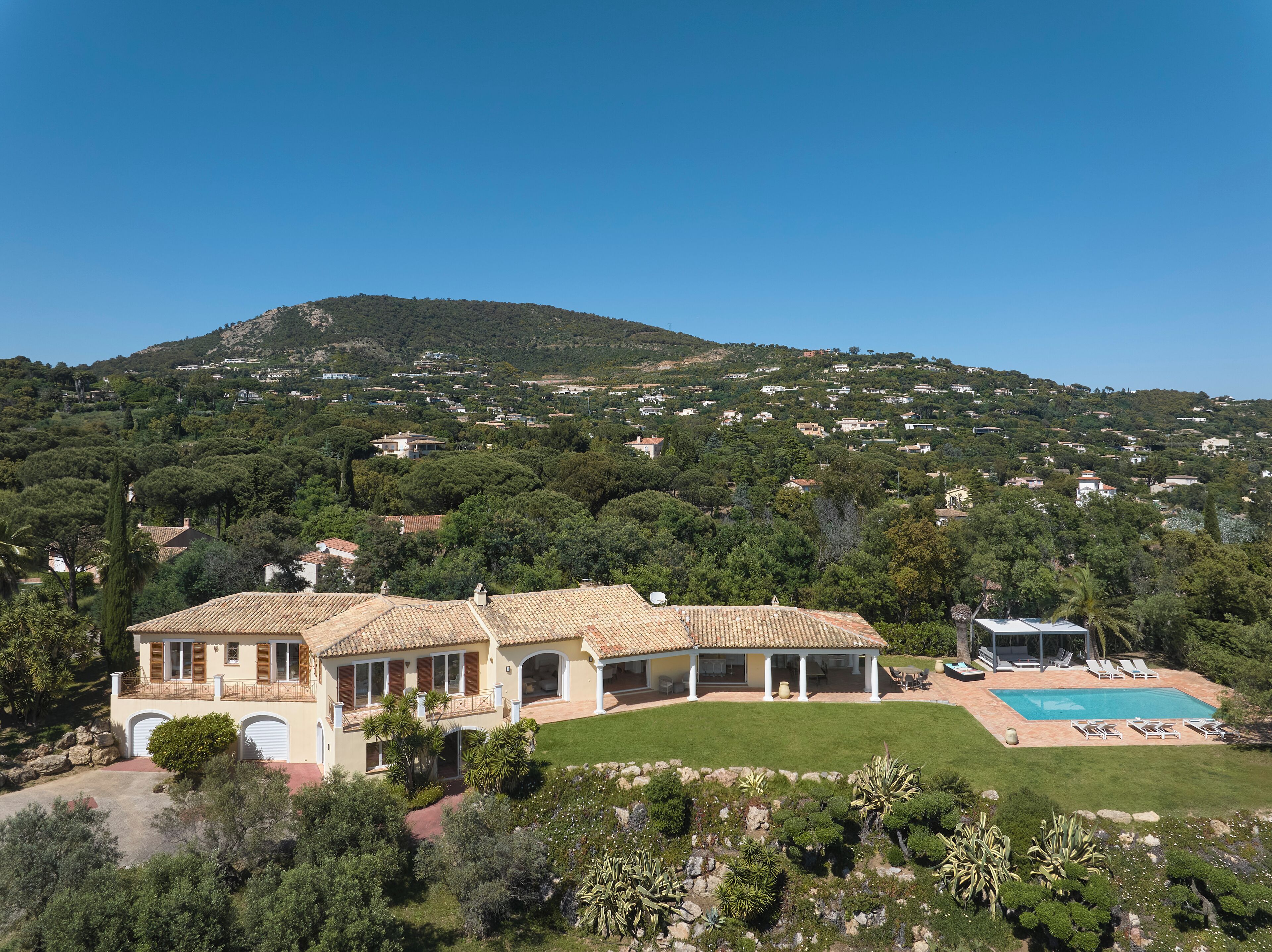 A Mediterranean-style villa with a swimming pool, surrounded by lush greenery and a hilly backdrop.