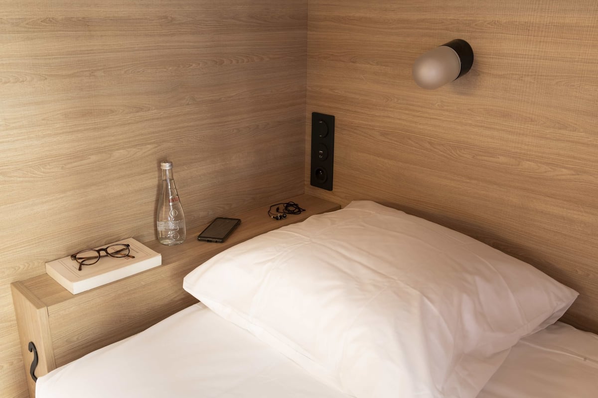 A neatly made bed with white sheets, a wooden nightstand with a bottle, glasses, and a smartphone.