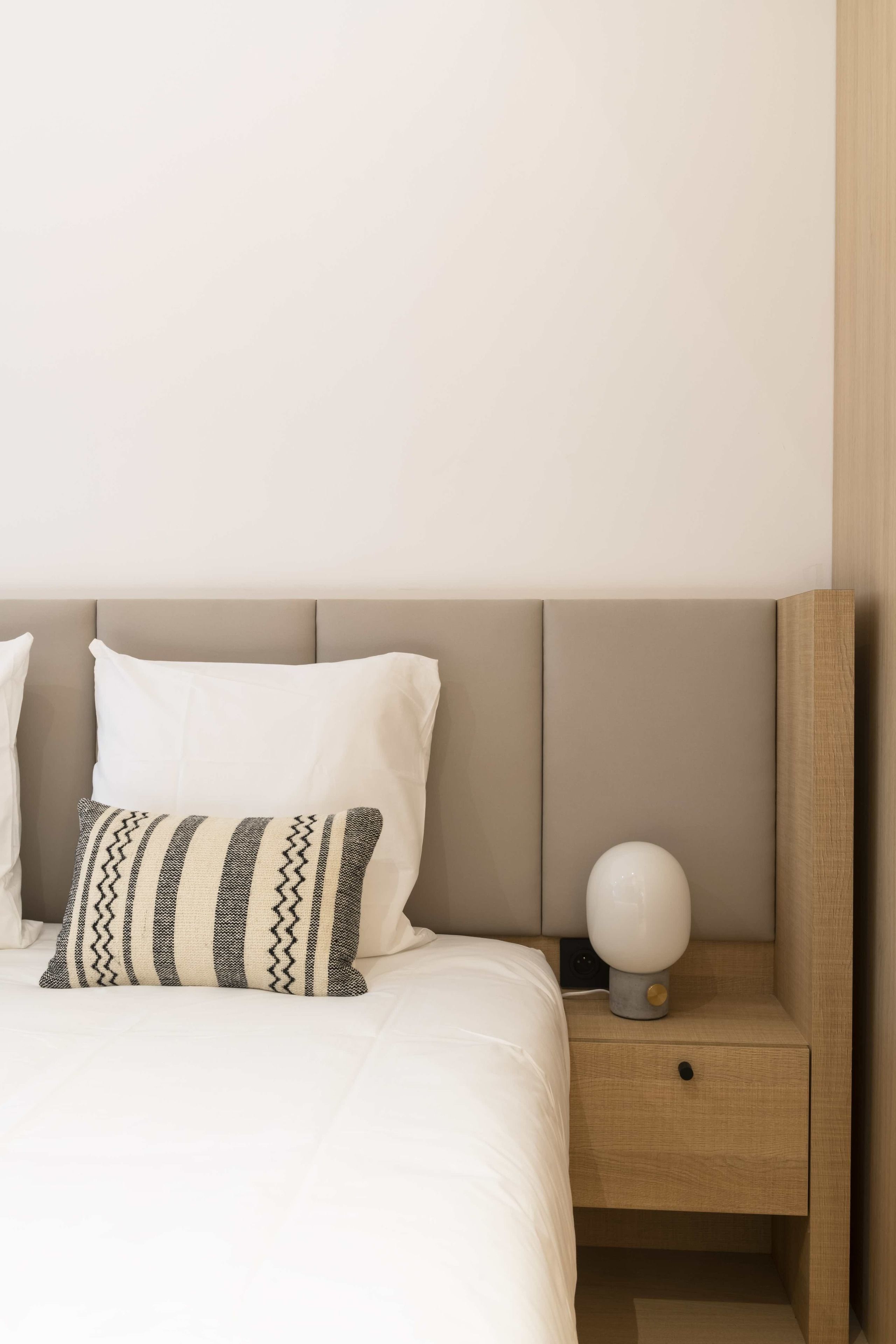 Una cama bien hecha con ropa blanca y una almohada a rayas, una mesita de noche con una lámpara y una mesita auxiliar de madera.