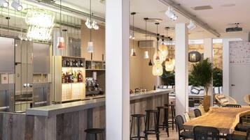 Un moderno bar con barra de madera, varios taburetes, lámparas colgantes y una zona de estar con una gran mesa de madera y sillas.