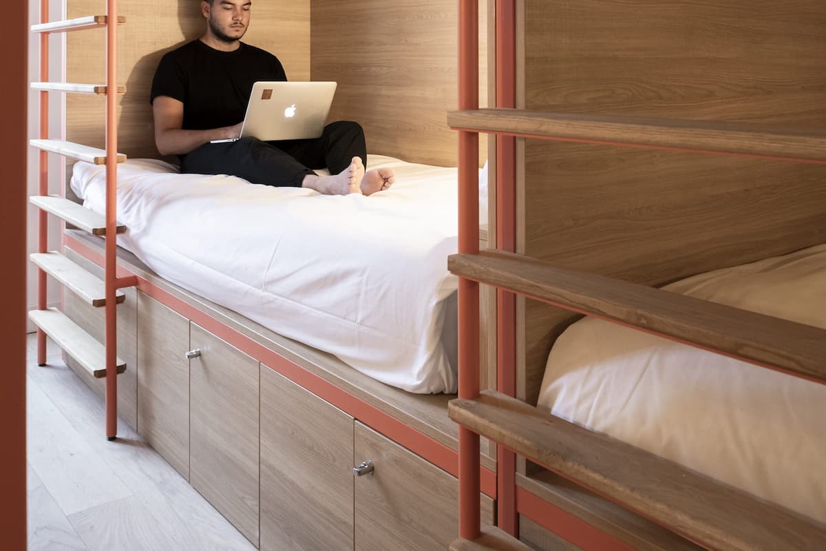 A bunk bed with a person using a laptop, wooden walls, and a metal ladder.