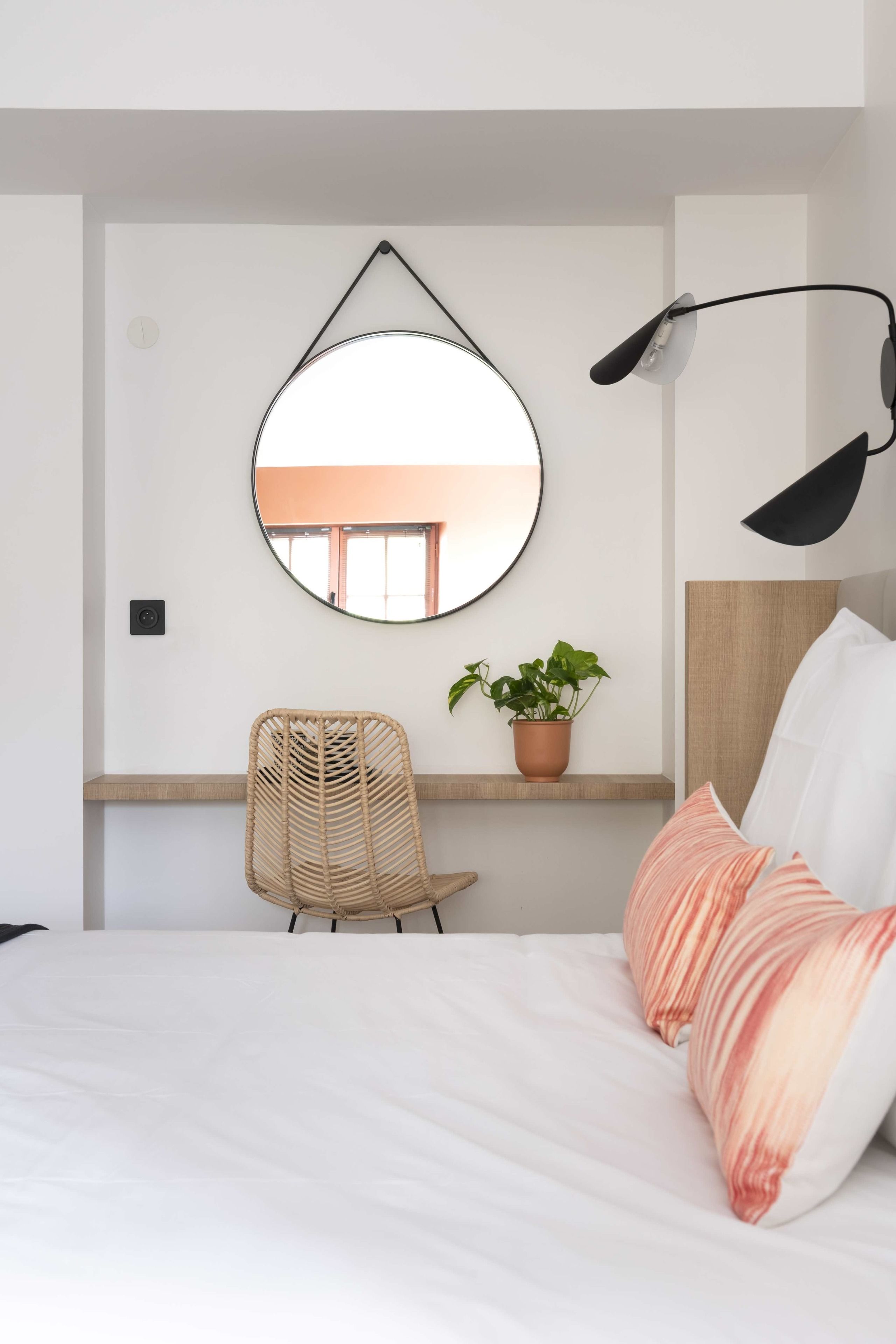 Ein Schlafzimmer mit einem Bett, einem runden Spiegel, einem Sessel, einer Topfpflanze und einem Schreibtisch mit einer Lampe.