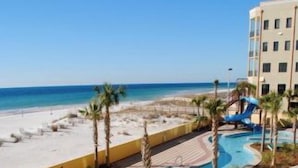 A swimming pool with a beach view, palm trees, and a building in the background. - The Oasis at Orange Beach  - Lazy River + Slide, Pools, Splash Pad, Cafe & Bar.. (Orange Beach)