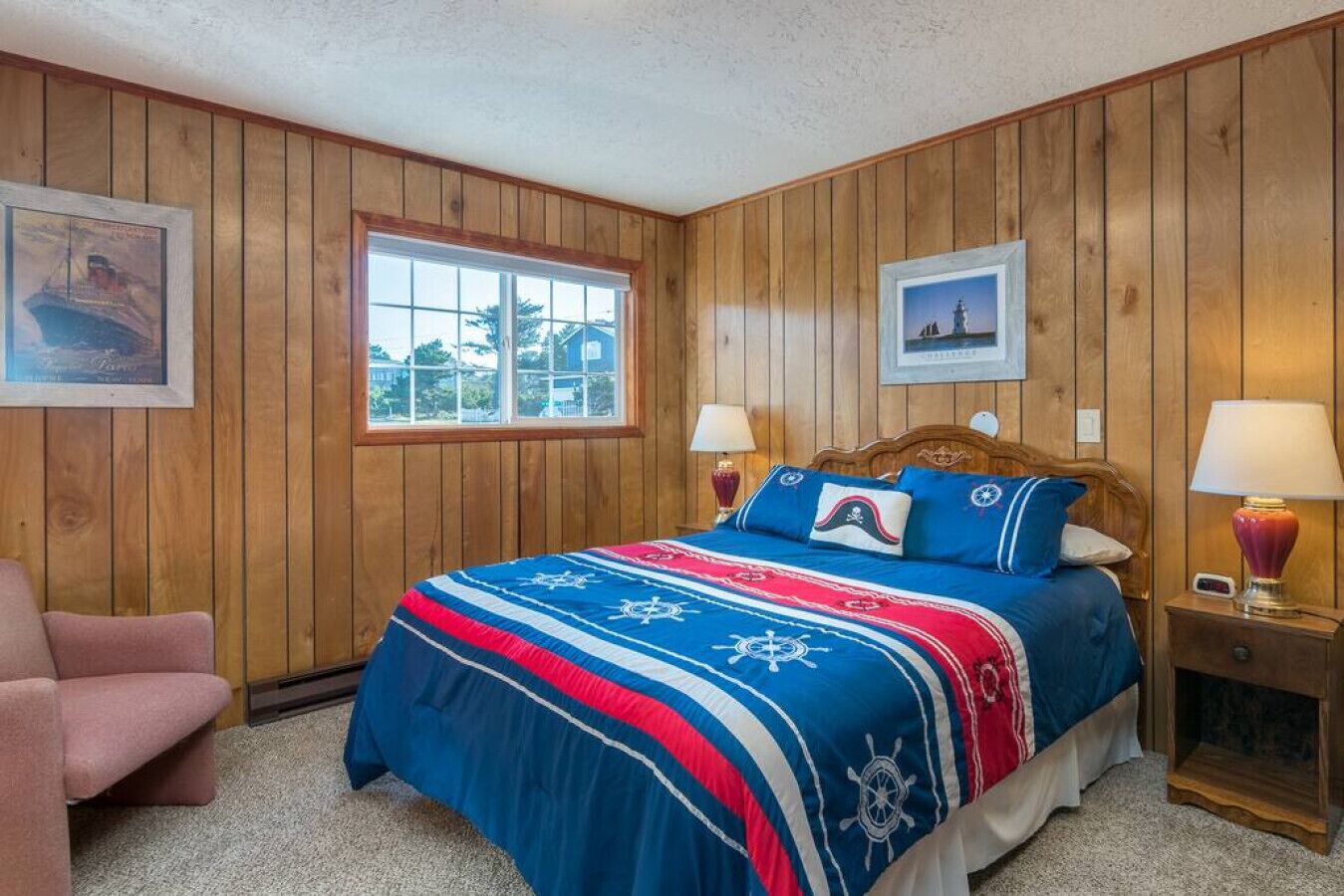 A bedroom with wooden paneling, a bed with a nautical-themed bedspread, two bedside lamps, and a window with a view of the outside.