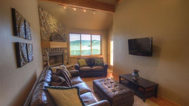 A living room with a stone fireplace, a large window, and a flat-screen TV mounted on the wall. - Smashing Location! - Affordable, Max. Amenities, Hot Tub, Views (Big Sky)