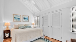 Une chambre à coucher comprenant un lit, deux tableaux accrochés au mur, une table de chevet avec une lampe, une fenêtre avec des stores et une porte à panneaux ajourés.