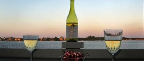 A table with two wine glasses and a bottle of wine, a bowl of fruit, and a view of the water and sky.