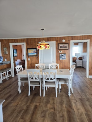 A dining area with a white table and chairs, a hanging light fixture, wooden panel walls, and a door leading to another room.