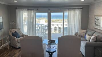 Una sala de estar con vista al océano, un balcón con mobiliario exterior y un comedor con dos sillas y una mesa.