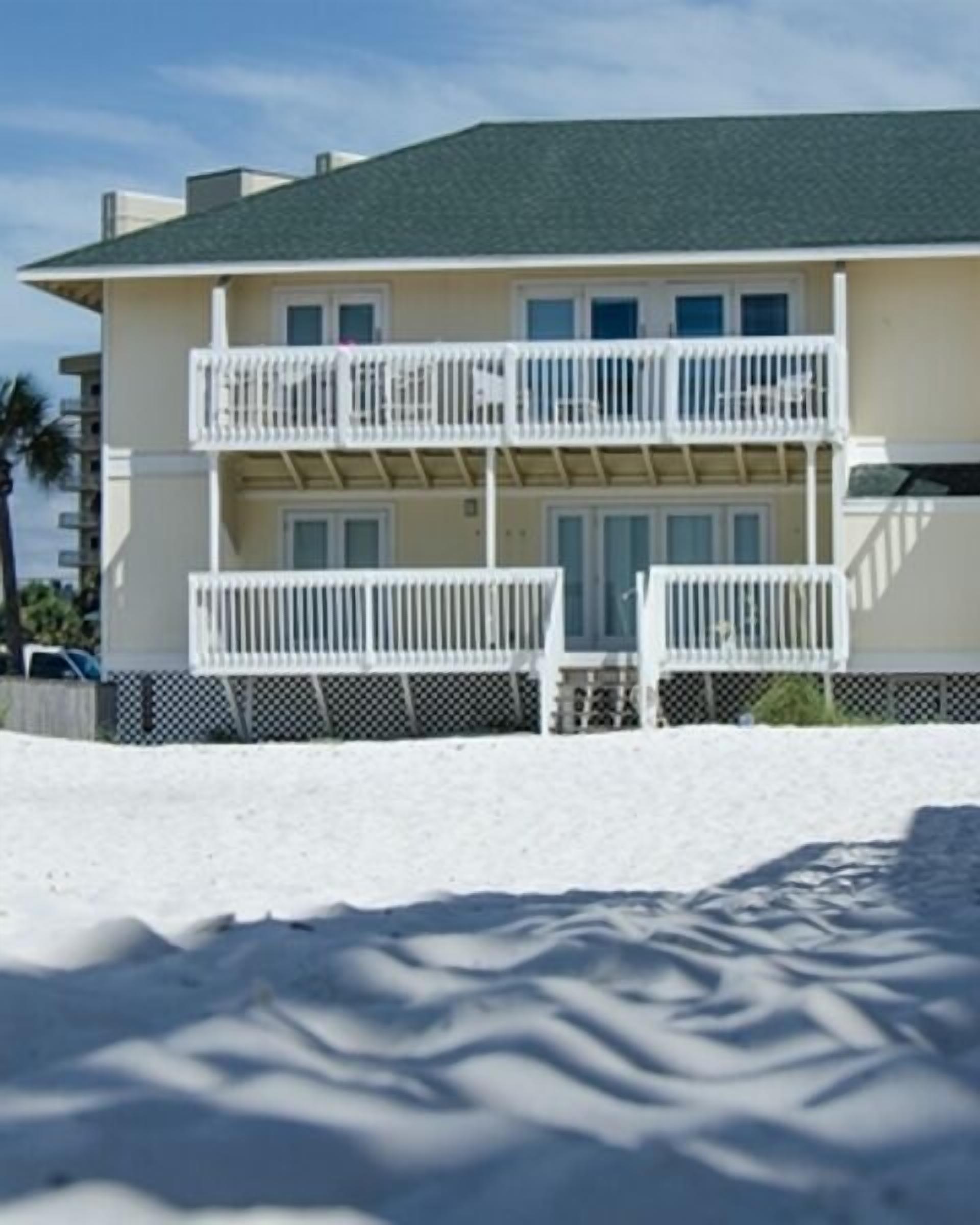 A two-story beachfront building with balconies, a sandy beach in front, and a clear sky.